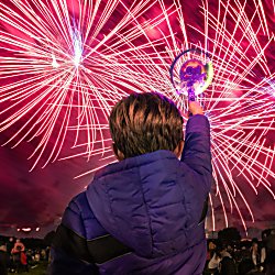 A boy holding up a toy light with fireworks being let off in the distance.