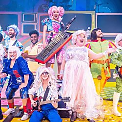 A cast from a panto on a stage at the Everyman Theatre. They are all dressed in bright coloured outfits with musical instruments.