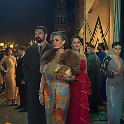 A photo of three people stood posing for a photograph dressed in old fashioned clothing. There are people behind them in groups too.