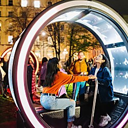 Two people sitting in a round light artwork looking at the roof of it. They are holding onto a bar in the middle of the artwork.