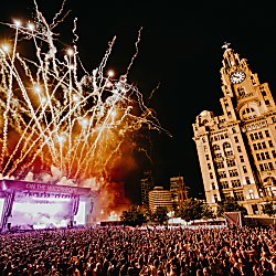 A stage with huge fireworks going off behind. There's a crowd of people watching the show at night time with the Royal Liver Building also in shot