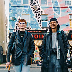 Two people wearing leather coats on Victoria Road in Wirral with a building covered in street art and a sign that reads 'rockpoint records'