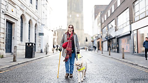 A guide dog user walking down Bold Street with her guide dog, the guide dog is wearing a harness and the guide down user has a walking stick in her right hand.