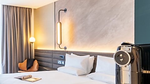 An extra large white bed in a spacious, airy room. The walls are a grey marble on one side and a deep green on the other. To the right is a special coffee machine and nearby a selection of coffee pods.