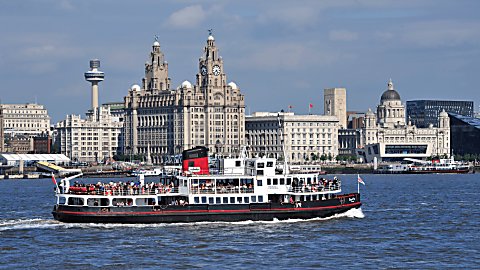 A white ferry on the River Mersey with the Liverpool skyline in the background. It features the Royal Liver Building, The Cunard Building and the Port of Liverpool building.