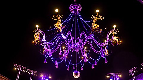 A large chandelier lit up in purple lights with performers on it as part of an act.