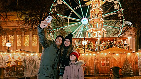 A family of three people, two adults an a child, taking a selfie in front of the Christmas Markets in Liverpool. St George's hall and a large ferris wheel is in the background.