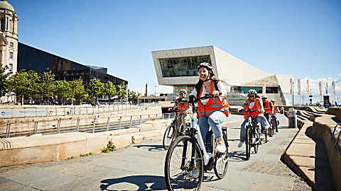 A group of people on electric bikes wearing orange high vis jackets on Liverpool's pier head.