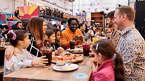 A family are sitting at bench tables in a food market. There are different coloured food vendor stalls in the food market.