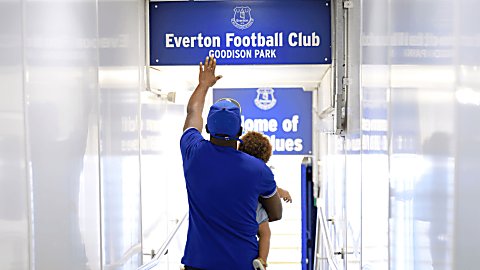 A person carrying a small child walking down the tunnel at Everton Stadium touching an Everton sign about the door.
