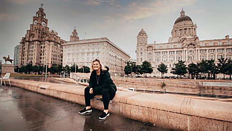 A person dressed in dark clothing sits on a wide stone ledge beside a wet waterfront promenade, with historic Liverpool buildings in the background, including the Royal Liver Building with clock towers on the left and a large domed building on the right. The ground appears rain-soaked, and the scene is lit by soft, overcast daylight with a muted sky.