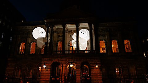 Two large glowing inflatable figures with cartoon-style eyes appear suspended between the columns of a grand historic building at night. The illuminated white sculptures contrast against the dark sky, while warm orange light shines through the building’s windows, creating a playful and dramatic light installation.