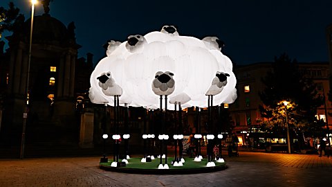 A large illuminated sculpture resembling a flock of sheep forms a glowing white cloud in a city square at night, with individual sheep heads and legs extending from the installation.