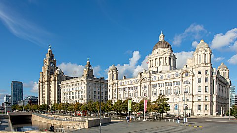 A wide daytime view of Liverpool’s waterfront featuring the Royal Liver Building with its clock tower on the left and the domed Port of Liverpool Building on the right. The historic white stone buildings line a tree-filled plaza with paved walkways, a few pedestrians, and flags on poles. The sky is bright blue with scattered clouds, and modern buildings are visible in the distance.