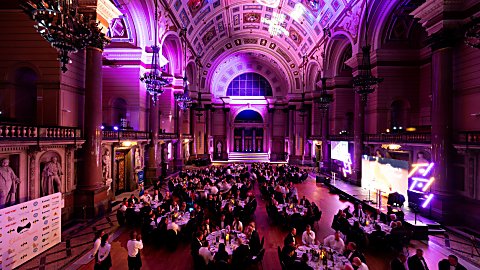 A grand hall with round tables and purple uplighters situated around the room with a stage to the right of the hall.