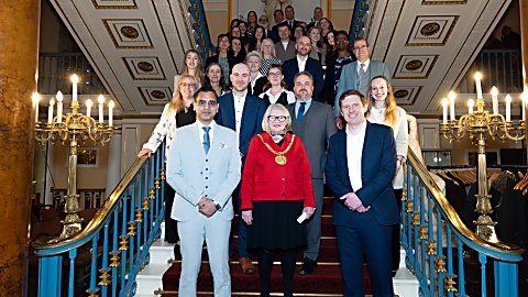 Large group of people standing on a grand staircase inside an ornate building, with chandeliers, decorative railings, and patterned carpet; one person at the front wears a ceremonial chain.