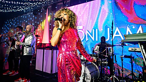 Live band performing on stage under string lights and colourful LED screens, with a lead singer in a bright pink sequined outfit singing into a microphone at centre, musicians playing brass, drums, and keyboards behind, and large screen graphics reading “Toni James” in the background.