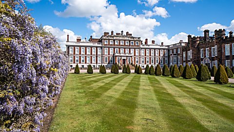 Wide view of a grand historic country house with red-brick and stone façades, set behind a neatly striped lawn and a row of conical trimmed trees, with purple wisteria in bloom along the left side and a bright blue sky with scattered clouds overhead.