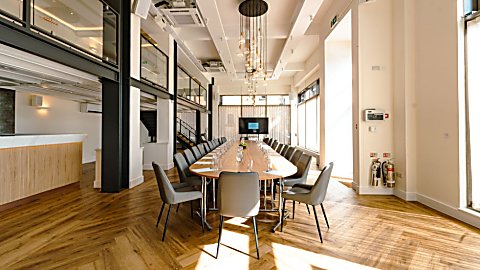 Bright, open-plan meeting and event space featuring a long boardroom table, wooden floors, high ceilings with modern pendant lights, large windows letting in natural light, and a presentation screen at the far end.