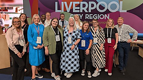 A group of people standing looking at the camera and smiling brightly. There is a sign behind them which says ''LIVERPOOL CITY REGION''.