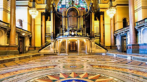 Close-up view of the revealed Minton tile floor in St George’s Hall, Liverpool, featuring intricate geometric patterns, colourful mosaic detailing and a central crest motif. Beyond the floor, a grand pipe organ stands beneath a stained-glass window, framed by sweeping staircases, ornate chandeliers, marble columns and richly decorated Victorian architecture illuminated by warm golden lighting.