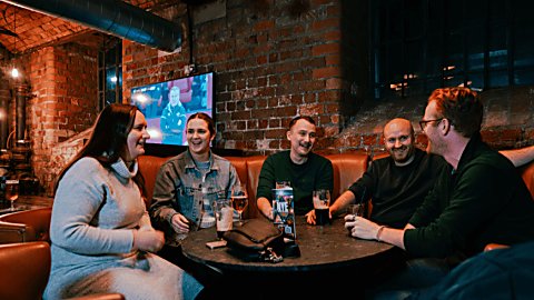 A group of five people sitting round a wooden table with a few drinks on the table. They are smiling and in conversation with a sports match on the tv in the background.