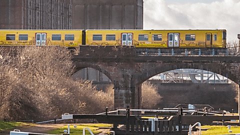 A yellow Merseyrail train passes over an old brick bridge which is over the canal and white locks. It is a sunny day and all the locks are closed. In the background a giant warehouse building can be seen.