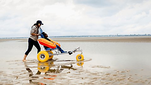 A person pushing an accessible wheelchair on a beach. The wheelchair has large yellow wheels and a wheelchair user is sitting in the chair enjoying the view of the beach.
