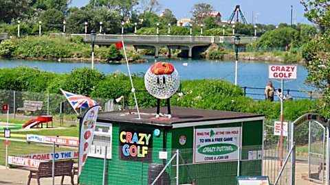 Crazy golf attraction beside a lakeside setting, featuring a green kiosk with colourful “Crazy Golf” signage and a large decorative golf ball wearing a crown on the roof. Mini-golf holes, flags, benches and fencing surround the area, with a bridge crossing the water and trees and greenery in the background. A sign reading “CRAZY GOLF” stands near the entrance.