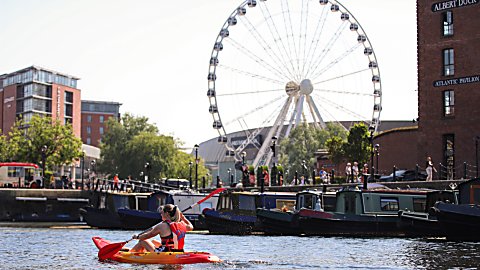 A person paddles a red and yellow kayak across the water at Liverpool’s Royal Albert Dock. Several narrowboats are moored along the waterfront, while the Liverpool Wheel rises prominently in the background. Historic brick dockside buildings, including one displaying signs for “Albert Dock” and “Atlantic Pavilion,” frame the scene on a bright sunny day.
