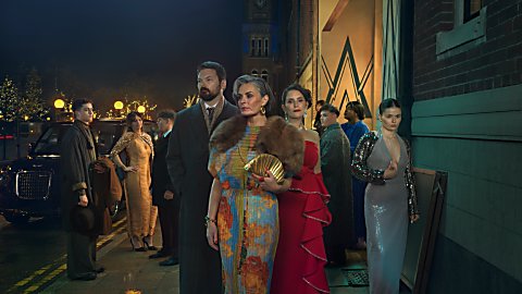 A photo of three people stood posing for a photograph dressed in old fashioned clothing. There are people behind them in groups too.