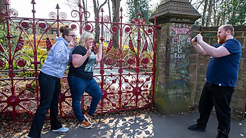 Two women smiling and posing while having their picture took by a man in-front of the Strawberry Field gates.