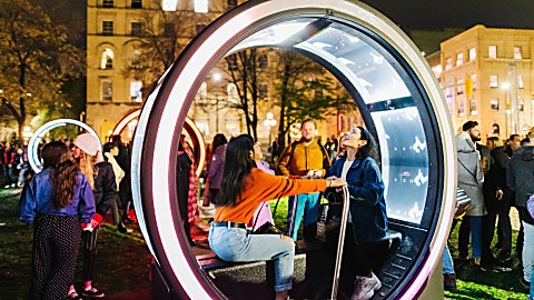 Two people sitting in a round light artwork looking at the roof of it. They are holding onto a bar in the middle of the artwork.