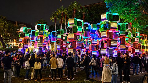 A light installation of stacked blocks in different colours with a crowd of people around it.