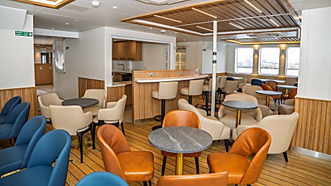 Modern indoor lounge on a ferry or ship with light wood flooring and wood-accented walls, featuring a central bar counter with high stools. Around the room are small round tables surrounded by cushioned armchairs in beige, blue, and tan. Large rectangular windows on the right provide a view of a city skyline over water. Overhead, slatted wooden ceiling panels and recessed lighting create a warm, contemporary atmosphere. Visible signage on the left indicates a muster station.