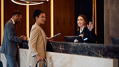 Two people standing at a reception desk with a receptionist standing behind the desk advising the person.