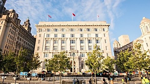 Cunard Building, the former headquarters of White Star Line in Liverpool, viewed from the waterfront. The historic stone building stands among the city’s Three Graces, with trees and public space in the foreground beneath a bright blue sky.