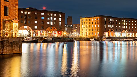 Night-time view of Liverpool’s Royal Albert Dock, with illuminated red-brick warehouse buildings reflected in the calm water. Lights from restaurants and waterfront businesses glow along the dockside, while boats are moored at the quayside and Liverpool Cathedral is visible in the distance against the dark evening sky.