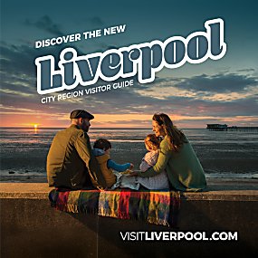 A family sitting at the side of a beach with two adults and two children cuddling together. The sun is setting in the distance and there is a long pier to the right of the image.