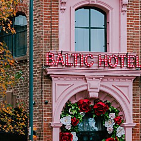 The exterior to an old warehouse building that has been turned into a hotel with a baby pink door front. The doorway has red and white roses hanging.