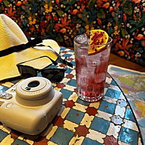 A polaroid camera with a cocktail and orange garnish, on the table is a hat and sunglasses.