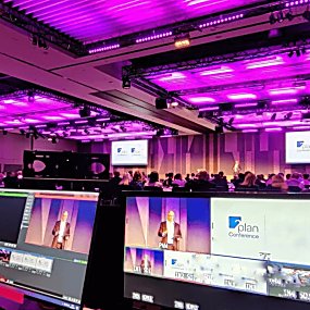 Inside a hotel conferencing room, the room is draped and lit with pink neon lights. There is a speaker on stage and the image is taken from the point of view of someone behind an AV desk with screens showing the event being broadcasted.
