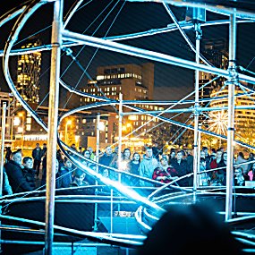 A huge metal structure that looks like a rollercoaster lit up in blue lights with crowds surrounding at night.