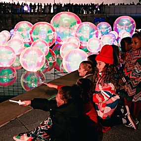 A selection of inflatable bubbles floating on a body of water that are lit up pink with a group of children watching.