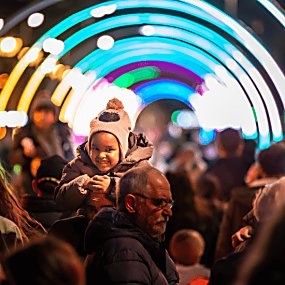 People are walking through a light tunnel in the dark. A young girl is smiling at the camera as she sits on the shoulders of a guardian.