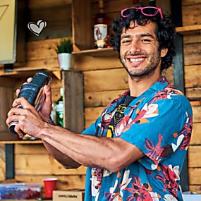 A man holding a silver cocktail shaker, smiles at the camera. He is wearing a blue and pink Hawaiian shirt and pink sunglasses on his head