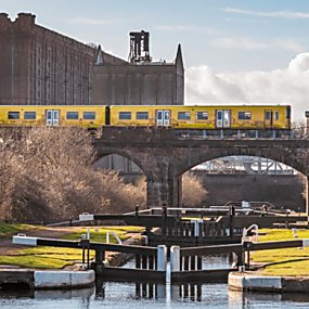A yellow Merseyrail train passes over an old brick bridge which is over the canal and white locks. It is a sunny day and all the locks are closed. In the background a giant warehouse building can be seen.