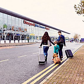 A man and woman are pulling small suitcase. There is a young child between them pulling a teddy bear suitcase. They are outside a modern, glass fronted building which is Liverpool Airport