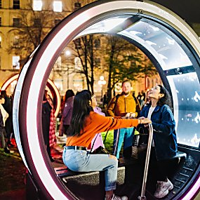 Two people sitting in a round light artwork looking at the roof of it. They are holding onto a bar in the middle of the artwork.