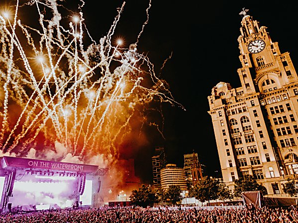 A stage with huge fireworks going off behind. There's a crowd of people watching the show at night time with the Royal Liver Building also in shot
